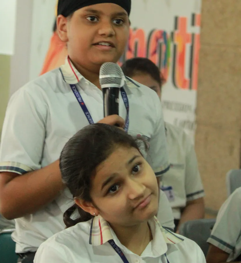 A schoolgirl giving a doubtful look while a classmate speaks into a microphone